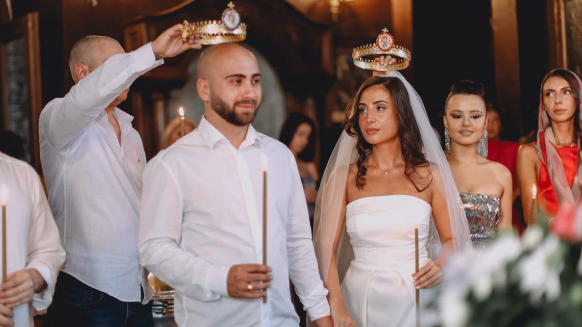 A wedding ceremony featuring a bride and groom holding candles, adorned with crowns, surrounded by guests.