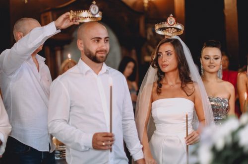 A wedding ceremony featuring a bride and groom holding candles, adorned with crowns, surrounded by guests.