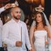A wedding ceremony featuring a bride and groom holding candles, adorned with crowns, surrounded by guests.
